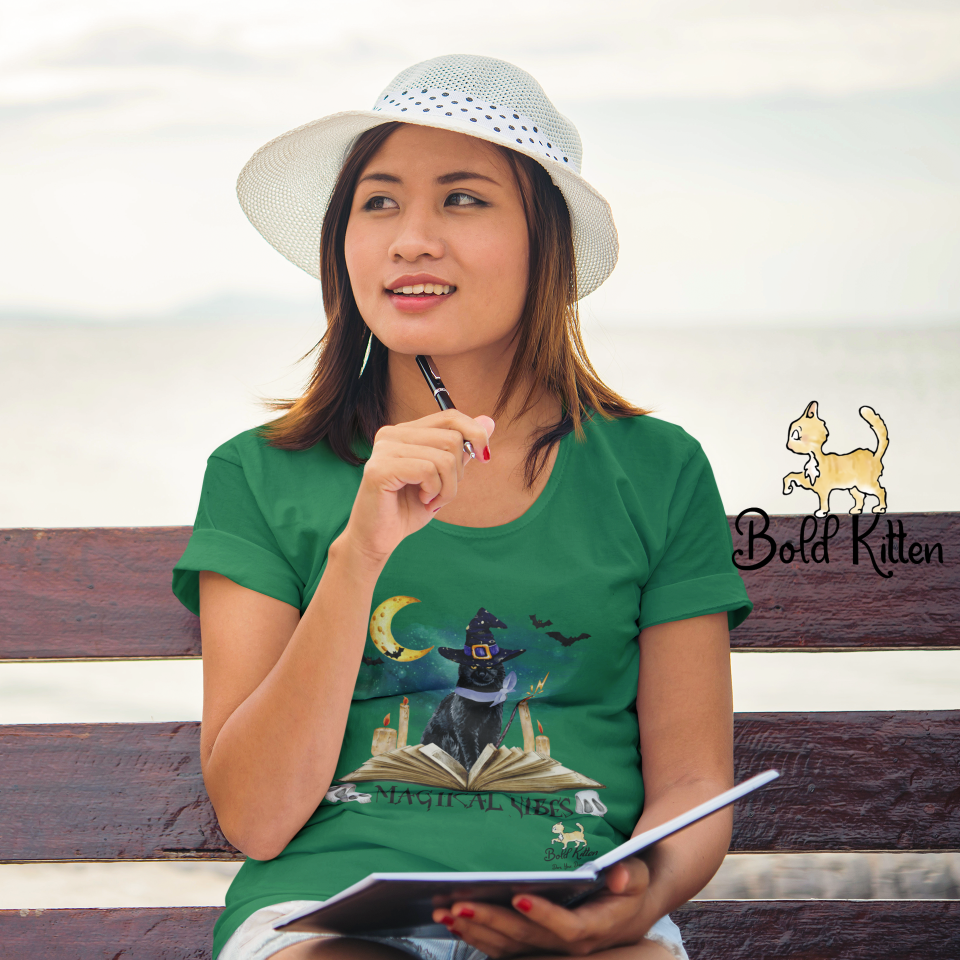 A young woman sitting on a bench near the sea holding a pen to her face, thinking, with an open notebook on her lap. She's wearing a green t-shirt with a picture of a cat wearing a witch hat sitting behind an open book. Below the book, bracketed by human skulls, are the words ‘Magikal Vibes’. In the background, bats fly and a yellow crescent moon shines.
