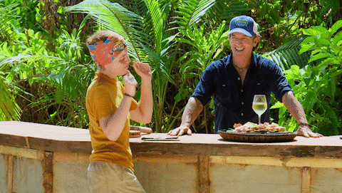 A person dancing next to a table with a glass of wine, in the jungle, behind which a person stands and watches 