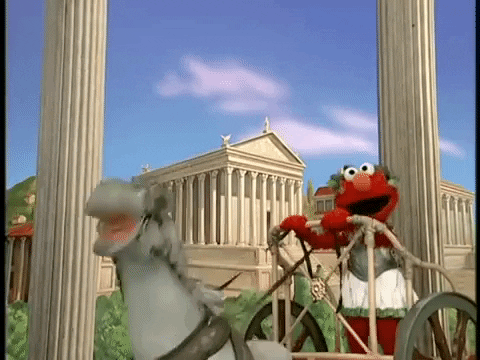 Elmo riding a chariot in front of ancient Roman buildings