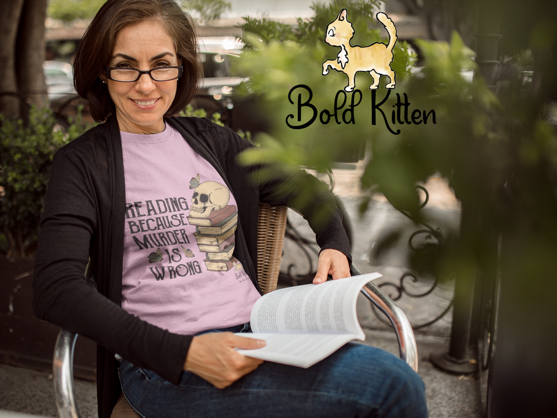 An older white woman wearing dark-framed glasses is reading a book while sitting at an outdoor table. She’s looked up from the book, which is currently resting on her blue-jean-clad legs. She is wearing a light pink shirt with ‘Reading because Murder is Wrong’ alongside a stack of books with a skull on top. There are butterflies on top of the skull and surrounding the word ‘is’. Greenery hangs low over the table, and among the leaves, Rocko, our Bold Circus Kitten, is lurking as only a Bold Kitten can.