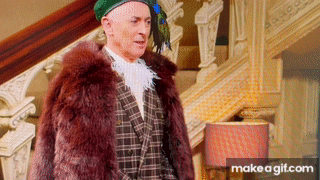 Alan Cumming, wearing a peacock feathered hat, turns and smiles at the camera