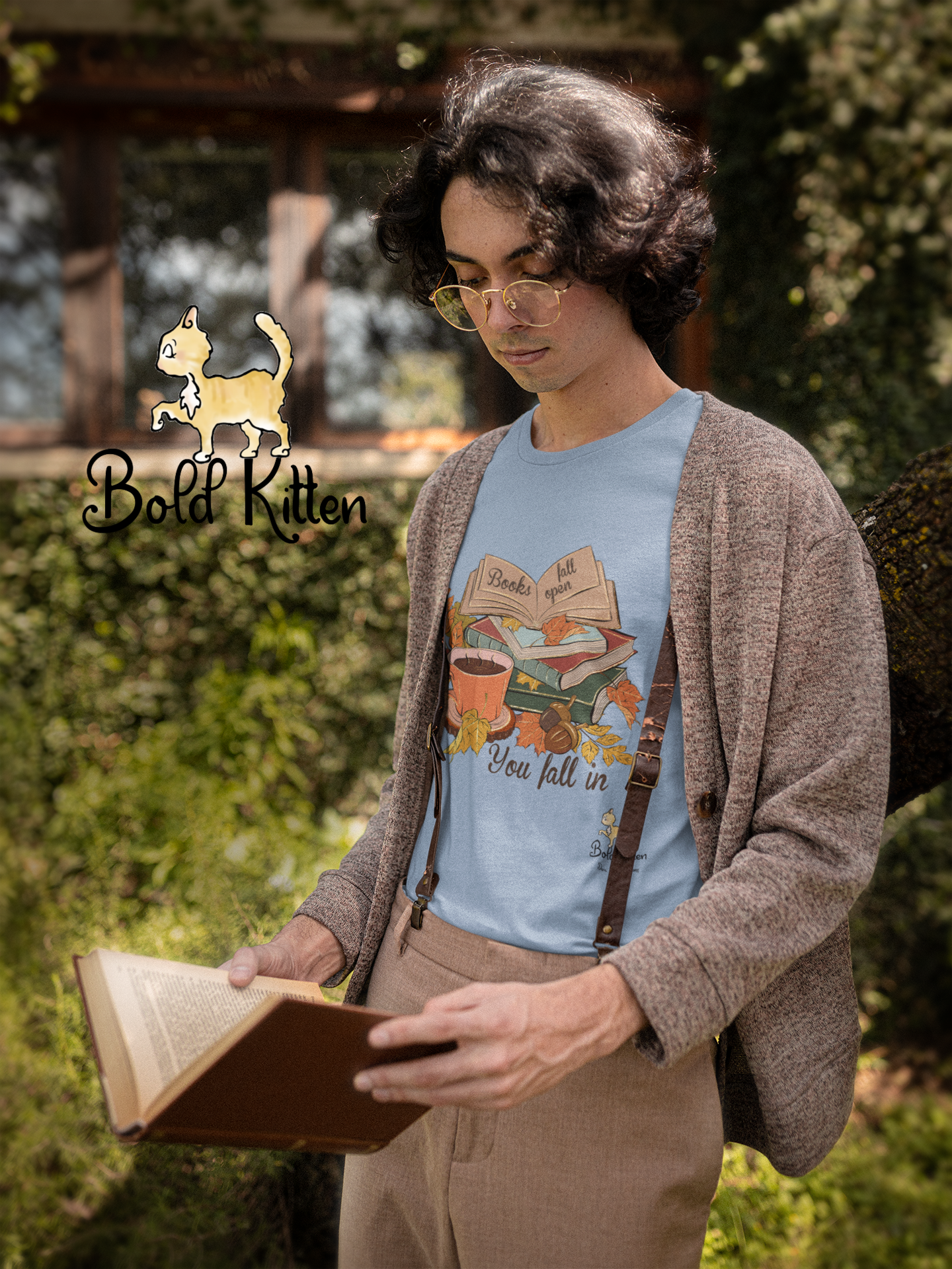 A young man with wavy black medium-length hair and gold wire-rimmed glasses is wearing a light-brown heathered sweater over a light blue t-shirt with a stack of books, some autumn leaves, and a cup of tea. The shirt reads ‘Books Fall Open, You Fall In’. Their light-tan pants are held up by a skinny pair of braces. As they stand there, they are reading a dark-red-bound hardcover book. Behind them is a riot of green – bush and vine. On top of the wall the vine is climbing, walks our Rocko, the Bold Circus Kitten, confident as ever as they stalk the brown brick.