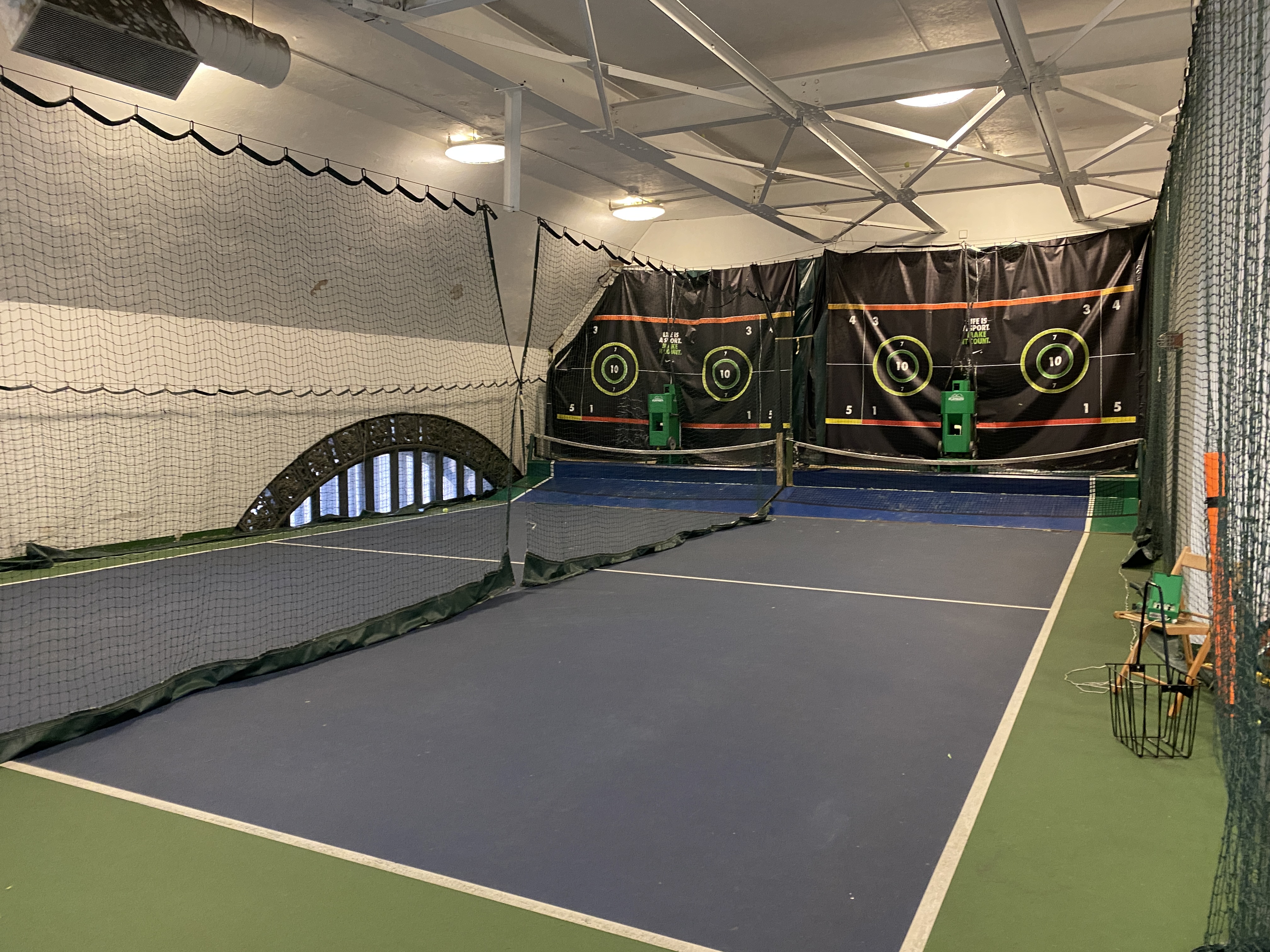 Two half-size tennis courts, indoors, separated by a net, with ball machines at the end