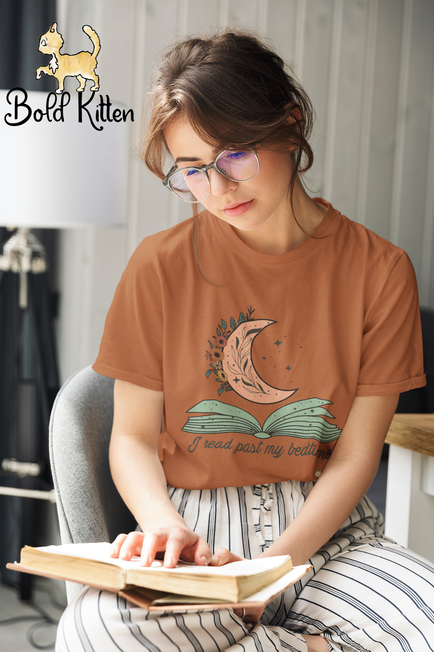 A young white woman with messily-styled short brown hair wearing large wire-frame glasses is sitting in a white modern-style chair with a desk to one side of her and a black-stemmed floor lamp (!) to the other side. On the top of the lampshade, Rocko the Bold Circus Kitten patrols the room, guarding the woman as she reads a book laying on her lap.   Her right leg is crooked on the chair. She’s wearing white pajama pants with thin black stripes and a brown-orange shirt with a pink-orange flowered half-moon with a few stars in the background over a greenish open book and the words ‘I Read Past My Bedtime’ in script below the book.