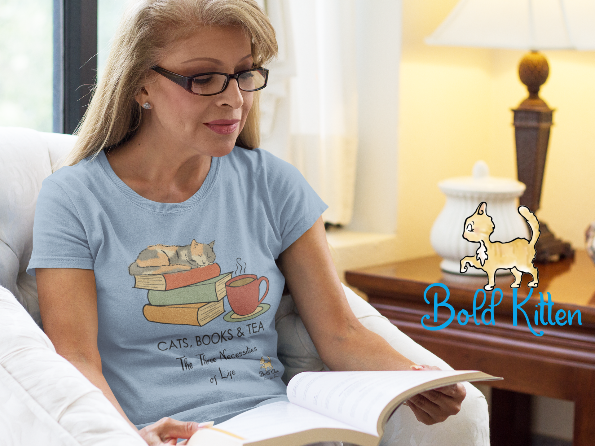 Middle-aged white woman with blond hair and dark glasses sitting in a comfortable chair, reading. They are wearing a light blue t-shirt that says 'Cats, Books, & Tea - the Three Necessities of Life'. Beside her is a lit lamp on a wooden table, On Which is a white ceramic jar and our Rocko, the Bold Circus Kitten.